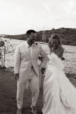 Black and white photo of laughing bride and groom walking by a sailboat on a tropical waterway.