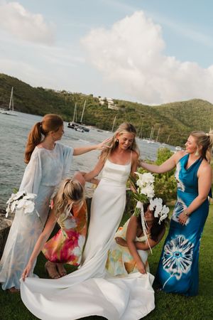 Bride in white gown surrounded by bridesmaids in colorful dresses with sailboats and green hills behind.