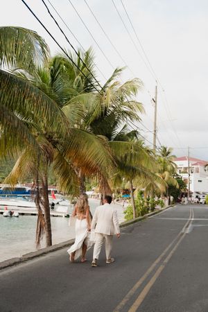 Newlywed couple walks hand-in-hand along tropical palm-lined street near marina in matching white wedding attire.