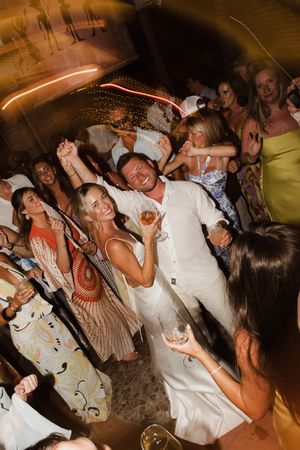 Couple in white outfits dancing and celebrating with guests at a lively wedding reception.