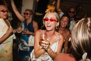 Joyful bride in white dress and red sunglasses laughing with champagne at lively wedding reception.