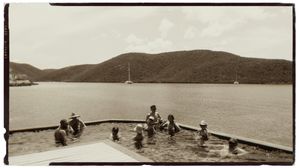 Vintage sepia photo of people swimming in an infinity pool overlooking a calm bay with sailboats and lush green hills.