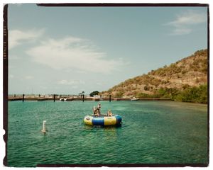 Two people on an inflatable water trampoline in a turquoise bay with a wooden dock and green hillside.