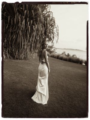 Bride in elegant backless white gown standing on lawn near tropical plants by the ocean.