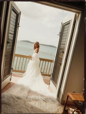 Bride in white ruffled wedding gown standing on balcony with ocean view through open doors.