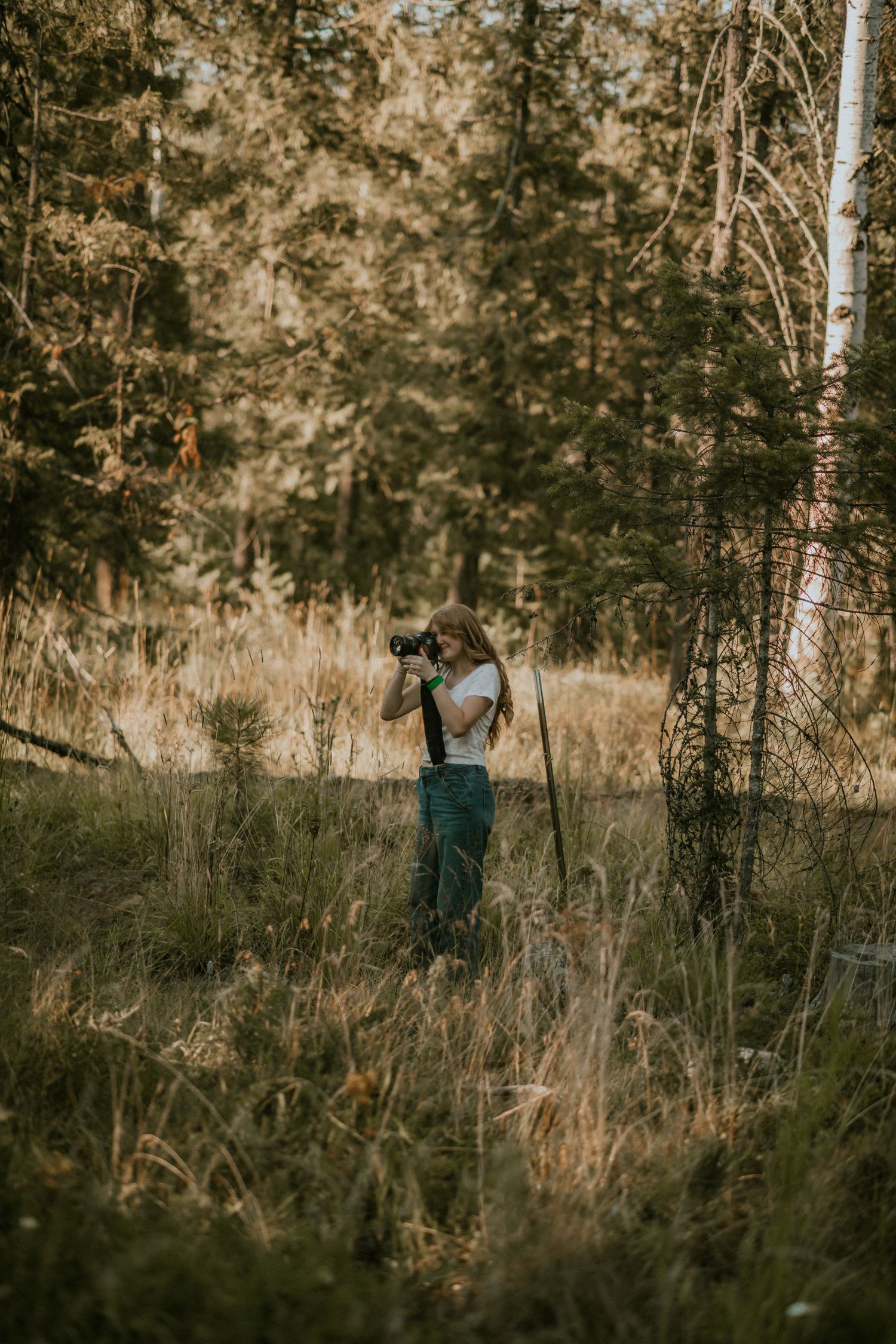 A woman with long hair photographs nature in a sunlit forest clearing, wearing green overalls and a white top.