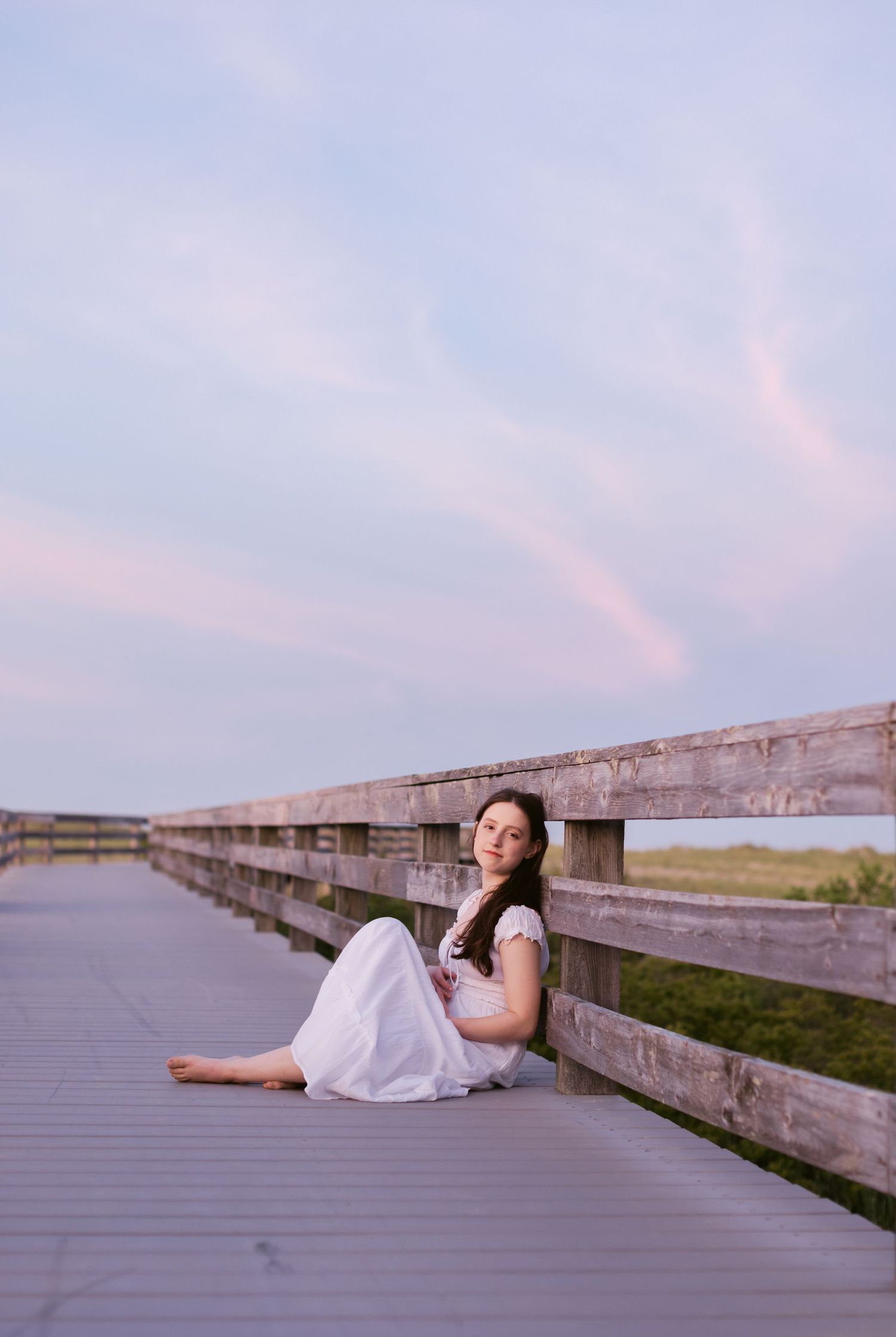 Senior photo shoot at Salisbury State Beach at sunset. Teen girl wears long white dress sitting on a gray boardwalk. Salisbury senior photos