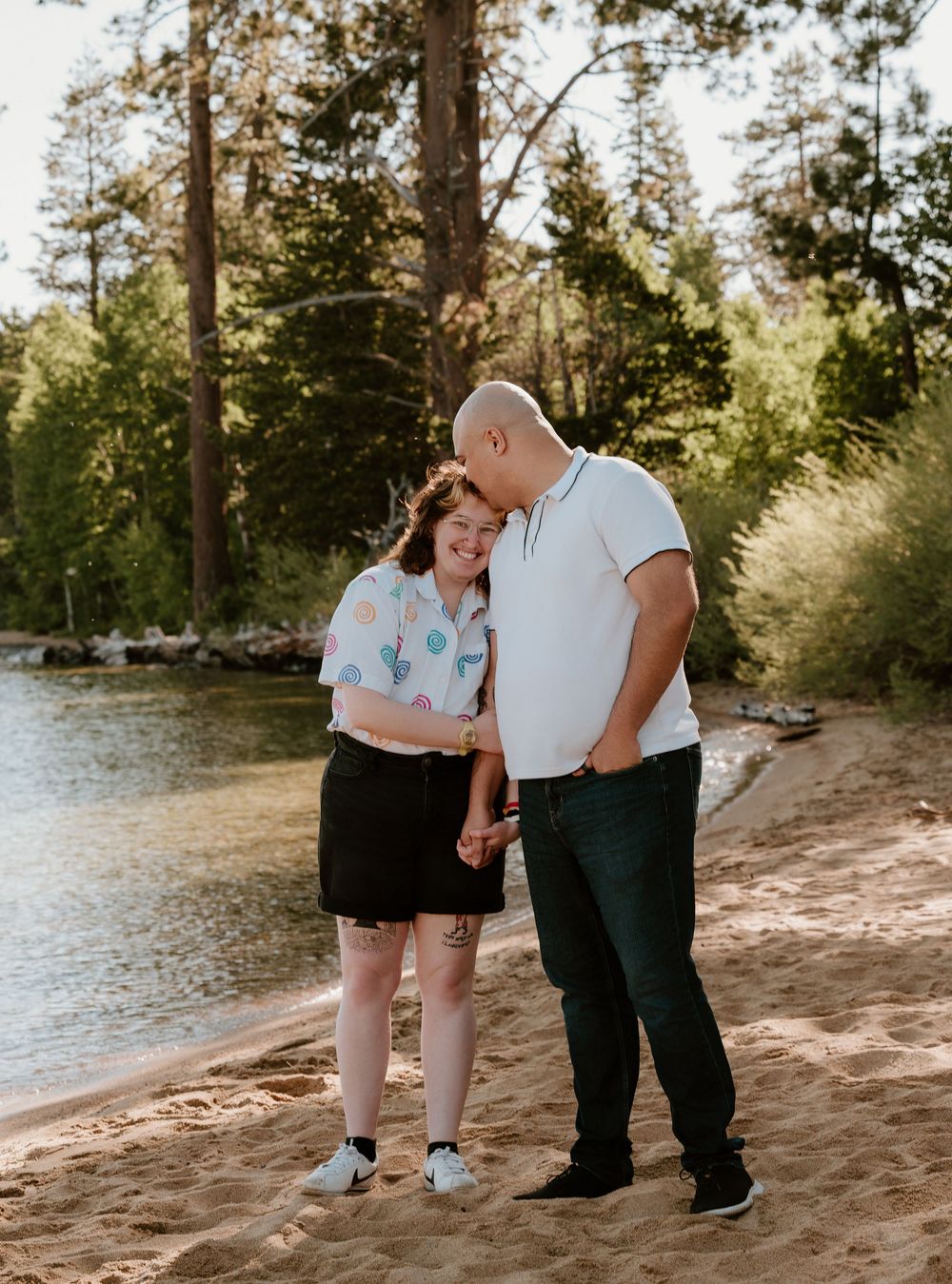 Happy couple holding hands on a sandy lakeside beach surrounded by tall pine trees in warm sunlight.