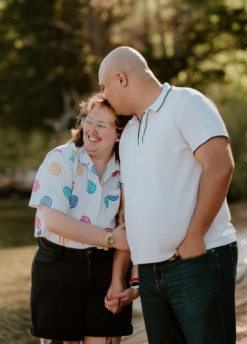 Couple holding hands and sharing a tender kiss outdoors, woman in colorful swirl shirt laughing joyfully.