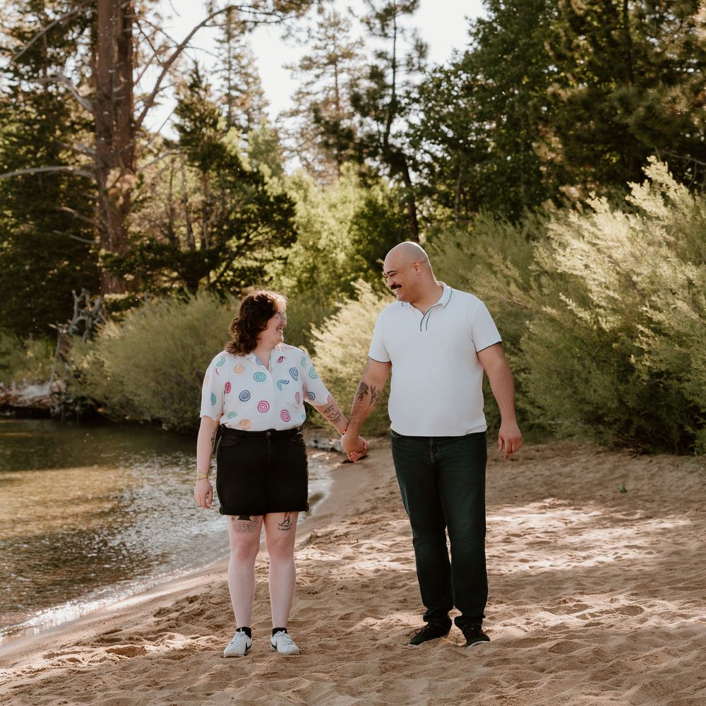 Happy couple walking hand in hand along a sandy lakeside beach with lush green trees behind them.