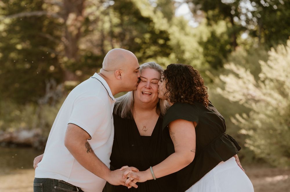 Joyful woman laughing as a bald man and curly-haired woman kiss her cheeks outdoors in warm light.