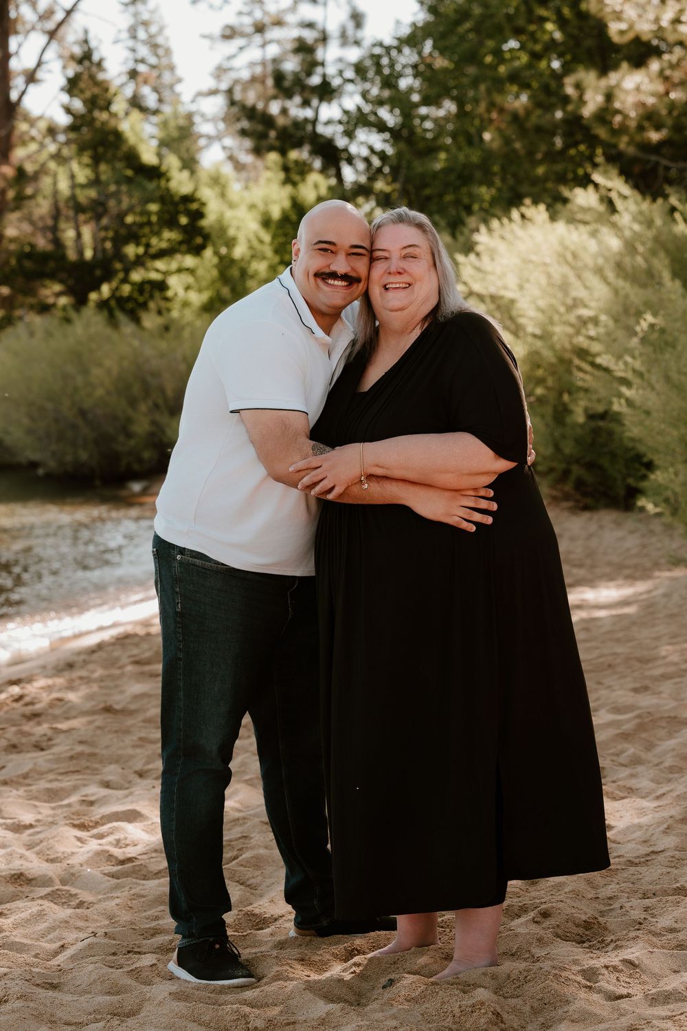 Couple embracing and smiling on sandy beach with lush green trees in background.