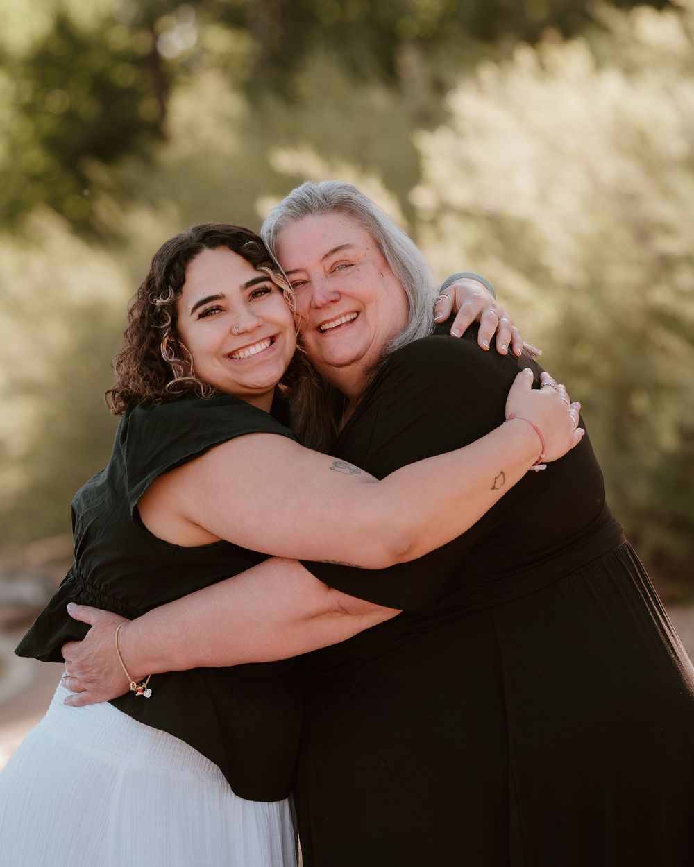 Two women sharing a warm embrace and smiling outdoors with soft green trees in the background.