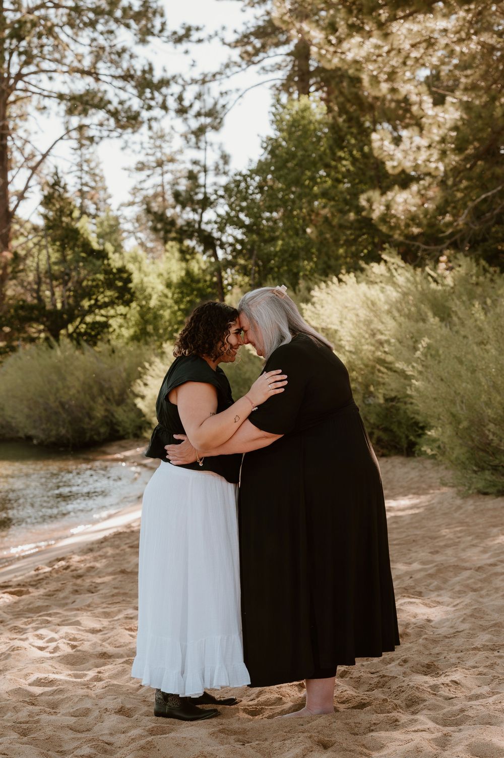 Two women in black and white dresses share a romantic embrace on a sandy beach surrounded by lush green trees.