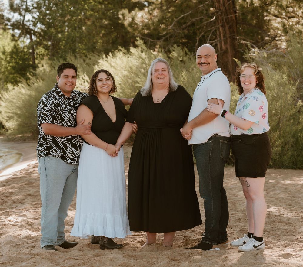 Five people smiling together outdoors on sandy ground surrounded by trees in a warm family portrait.