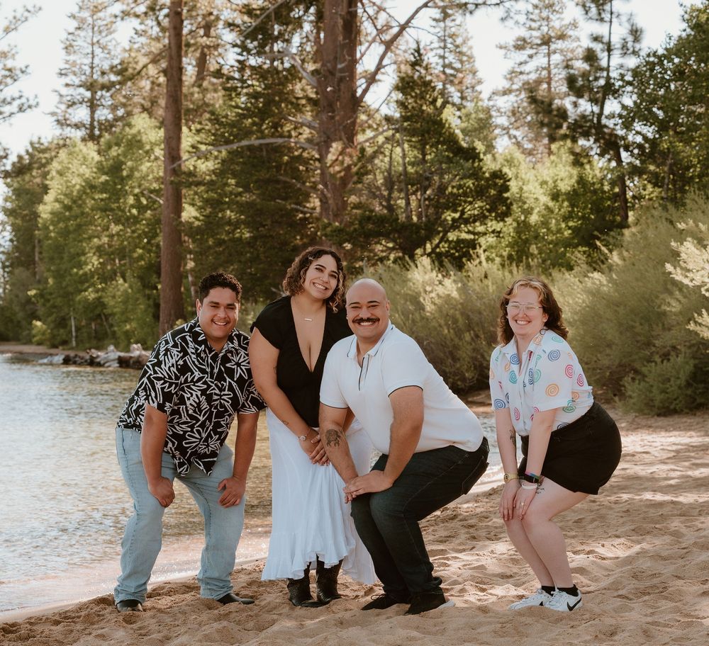 Family of four posing playfully on a sandy beach surrounded by tall pine trees near a lake.