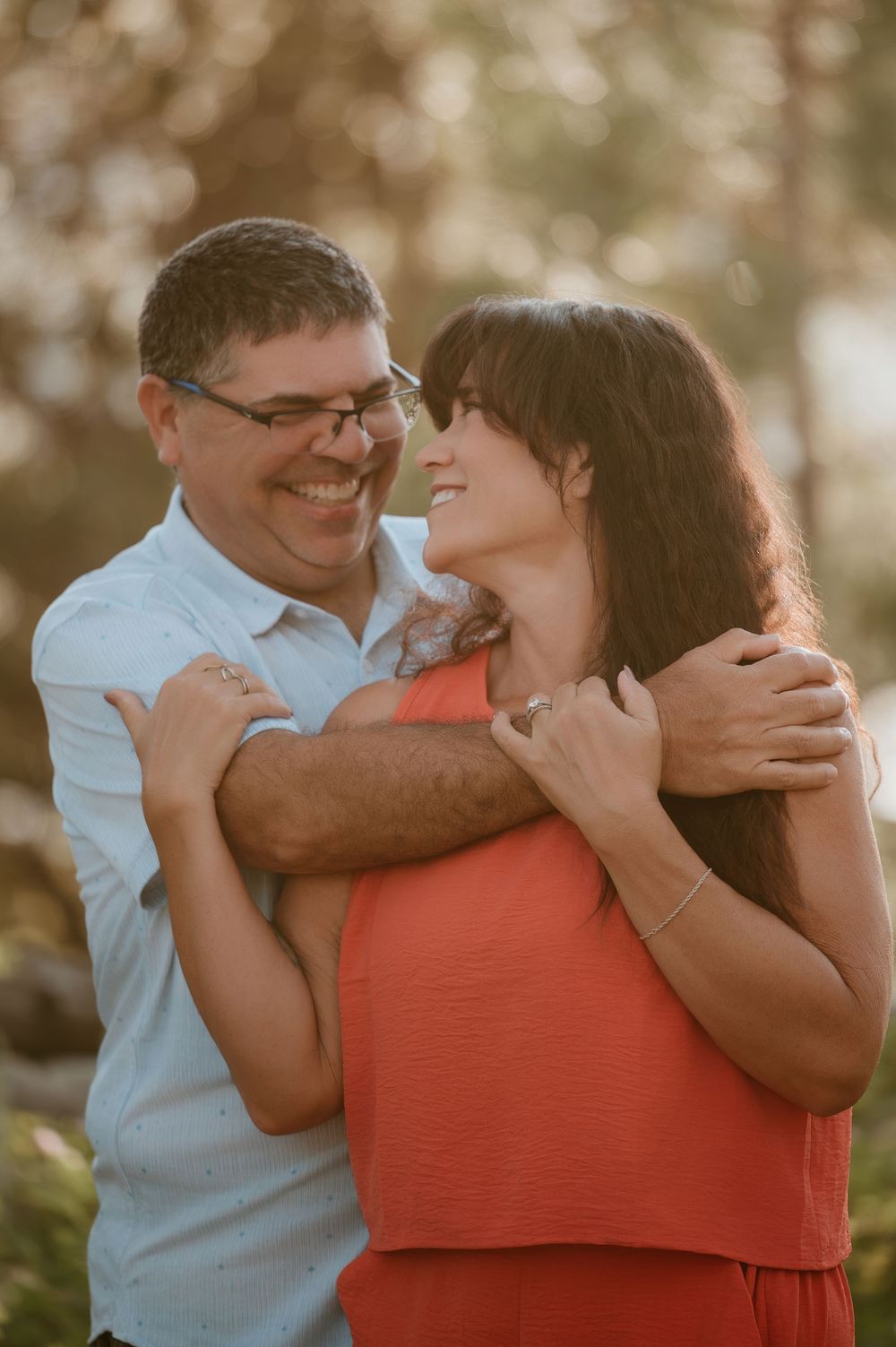Smiling couple embracing outdoors in warm golden sunlight, man in blue shirt hugging woman in coral dress.