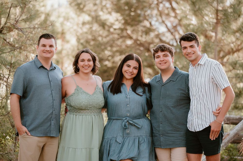 A smiling family of five poses outdoors among trees in casual summer attire for a warm family portrait.