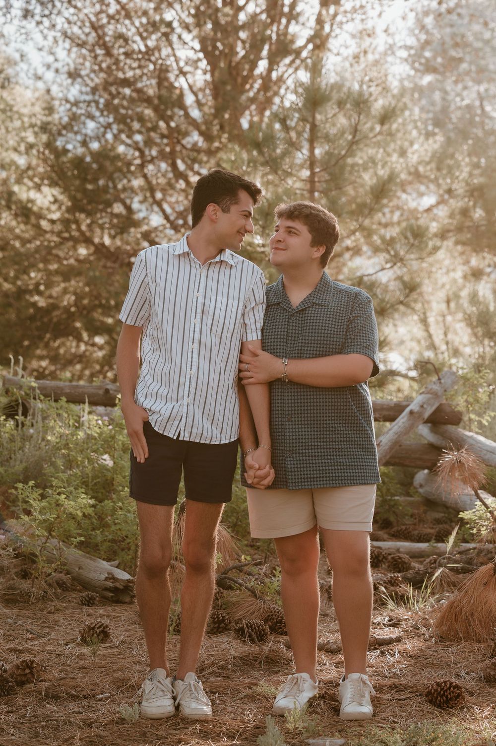 Two young men holding hands and smiling at each other outdoors among pine trees and natural scenery.