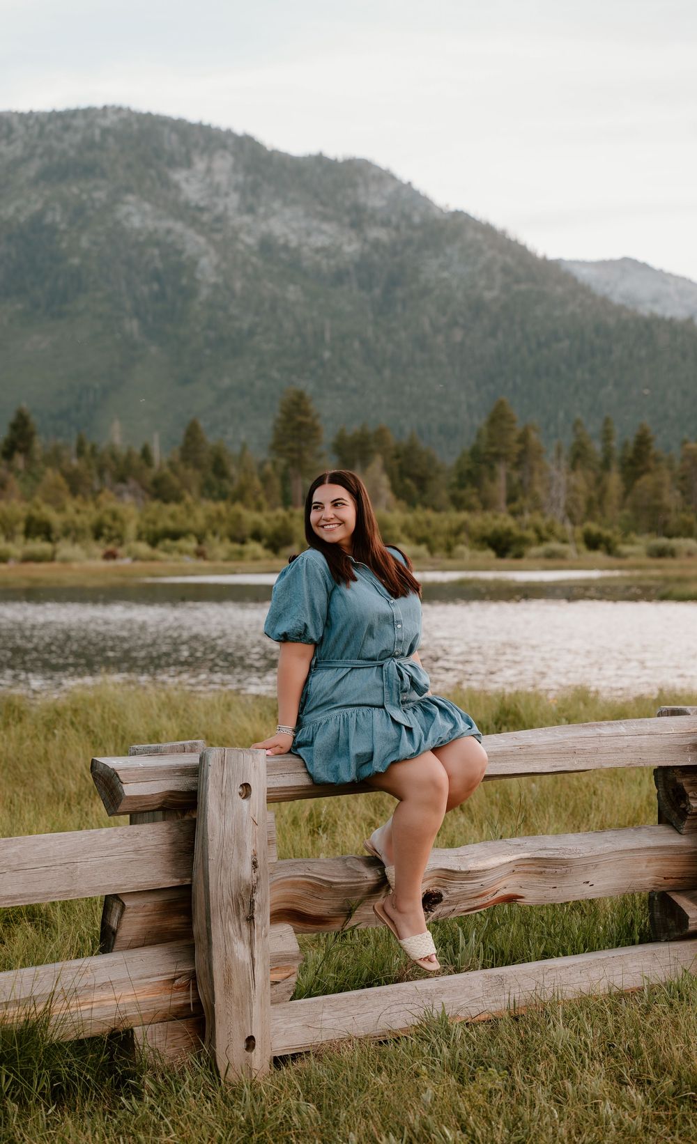 Young woman in teal dress sitting on wooden fence with mountain lake scenery behind her.