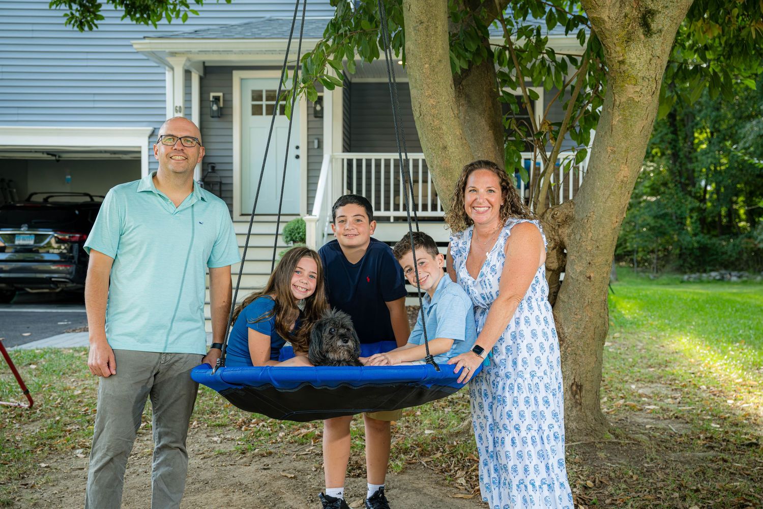Family of five smiling outdoors near a tree swing in a suburban backyard on a sunny day.