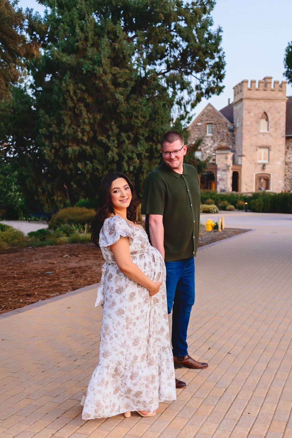 Pregnant woman in floral dress and man in green shirt pose on a pathway in front of a stone castle building.