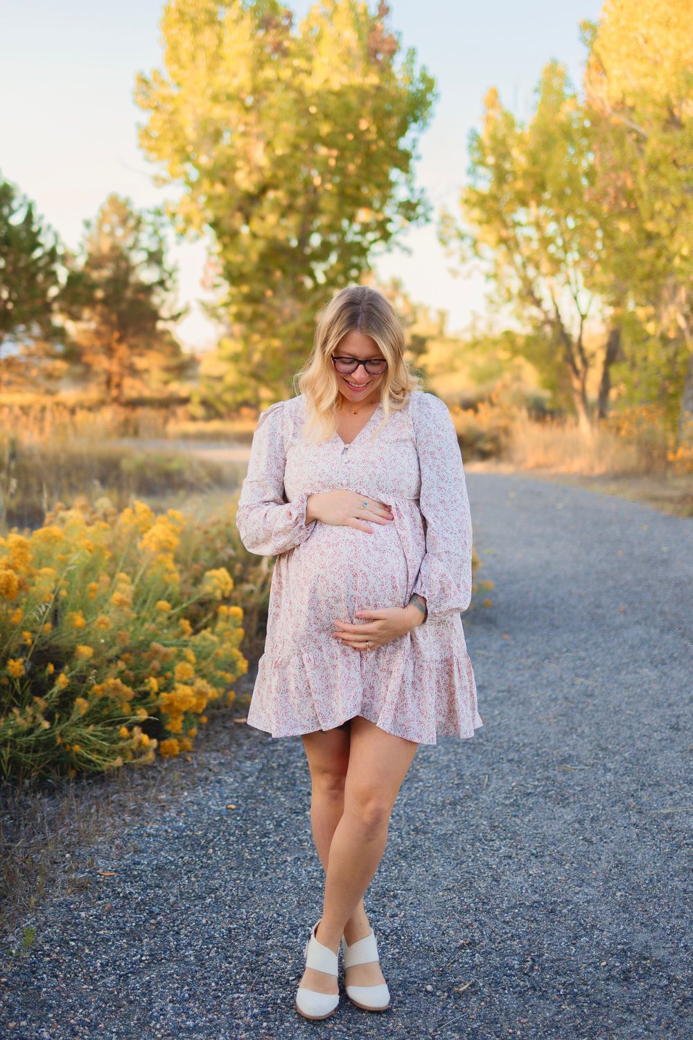 Pregnant woman in floral dress cradling baby bump on gravel path surrounded by golden trees and yellow wildflowers.