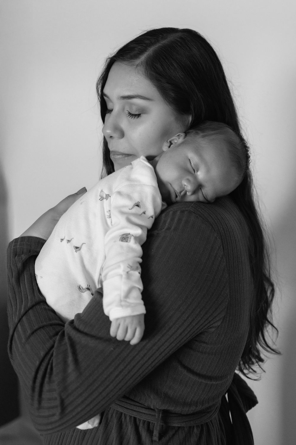 Black and white photo of a young mother with long dark hair gently kissing her sleeping newborn baby.