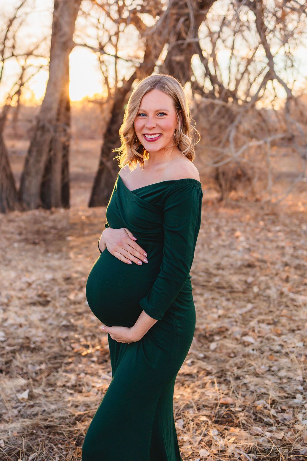 Smiling pregnant woman in green off-shoulder gown cradling baby bump outdoors at golden hour.