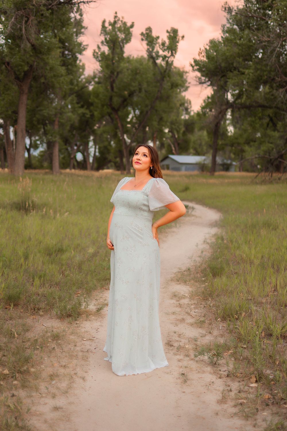 Pregnant woman in elegant white gown standing on dirt path surrounded by lush green trees at sunset.