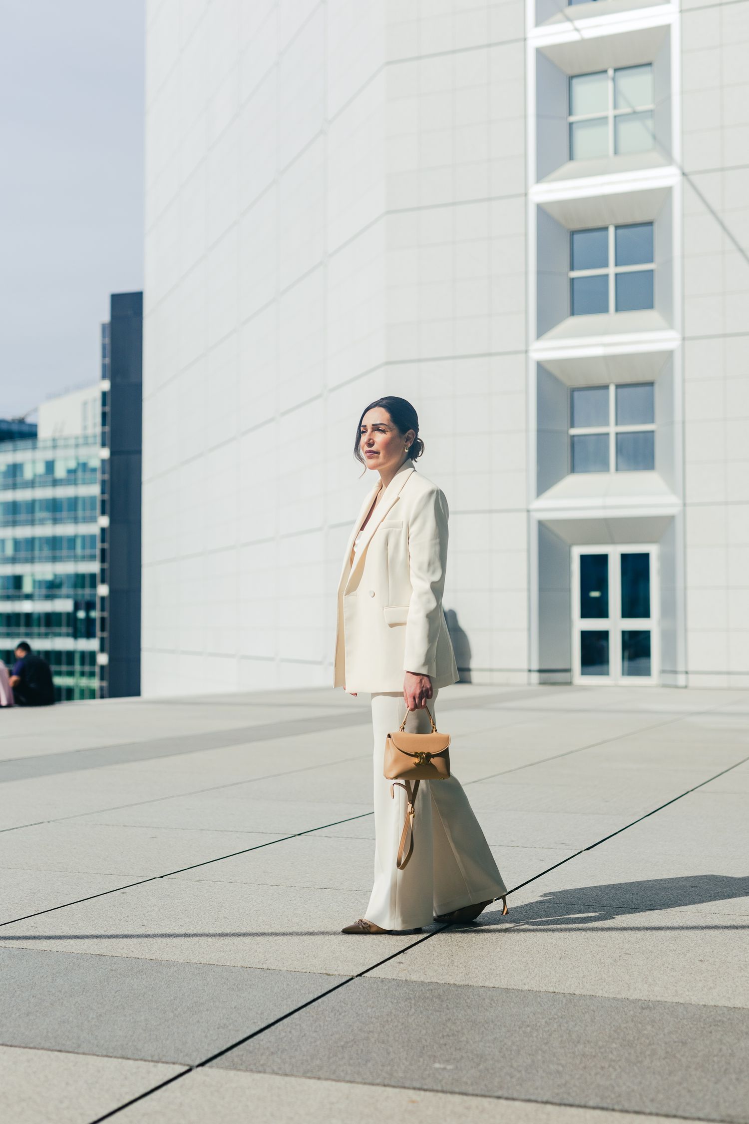 A stylish woman in a white pantsuit holding a tan bag walks confidently in a modern urban plaza.