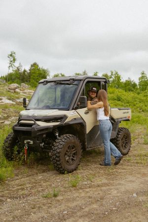 Woman standing beside a Can-Am UTV off-road vehicle on a dirt trail surrounded by green wilderness.