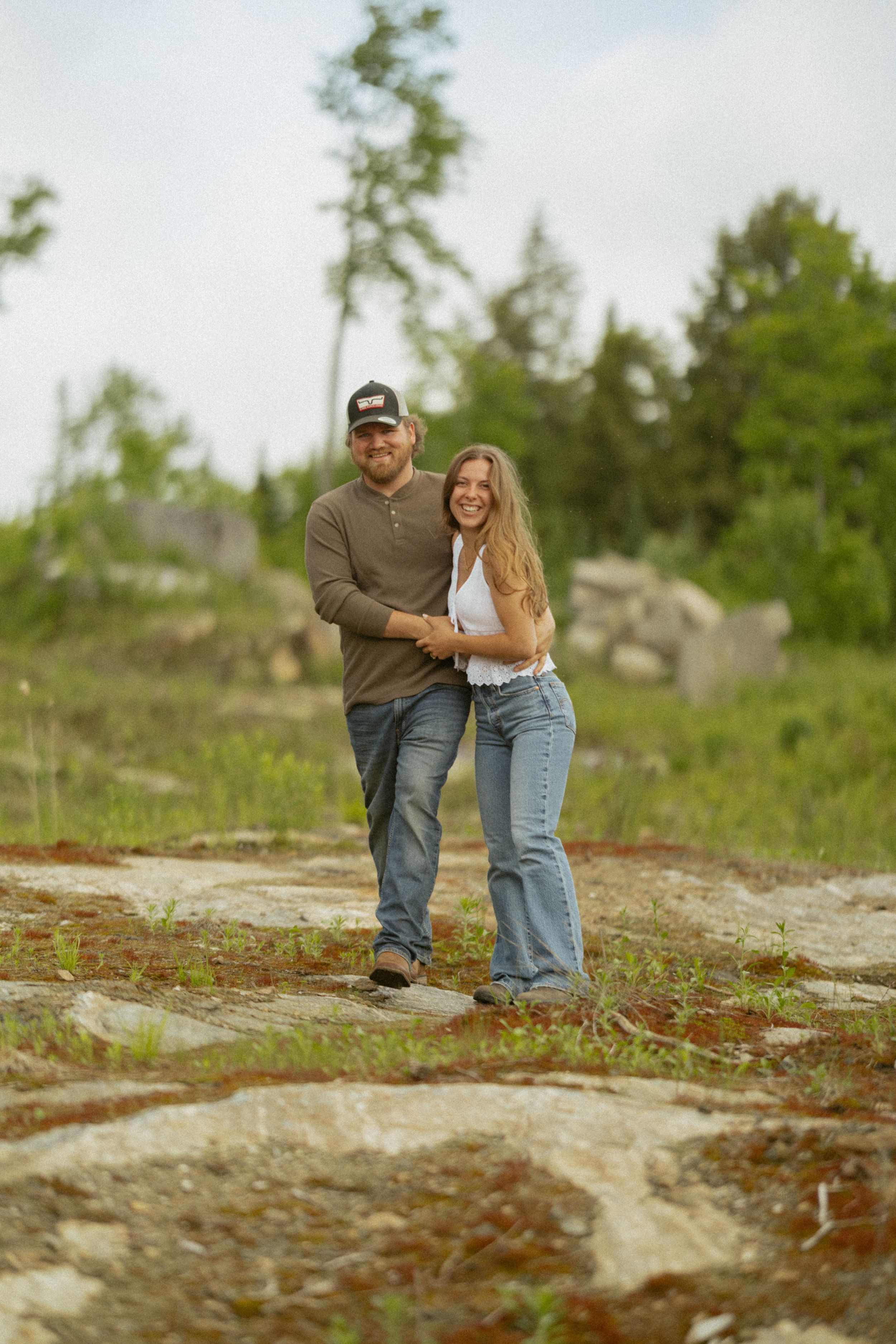 A smiling couple embraces outdoors on rocky terrain with lush green trees in the background.
