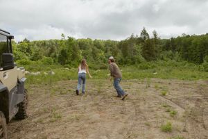 Two people walking toward a lush green forest in a rural clearing next to an off-road vehicle.