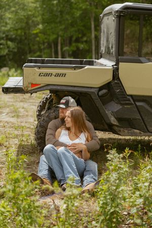 Couple sitting together in front of a Can-Am UTV in a wooded outdoor setting.