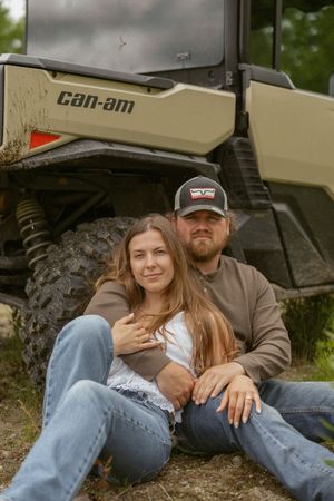 A couple sits together in front of a muddy Can-Am UTV in a wooded outdoor setting.
