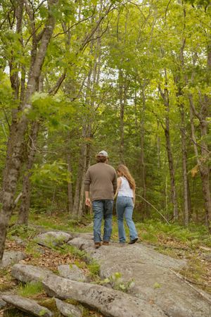 Couple walking hand in hand on a rocky forest trail surrounded by lush green trees in autumn.