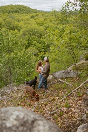 Couple sharing a romantic moment on a rocky hillside trail surrounded by lush green forest with two dogs nearby.