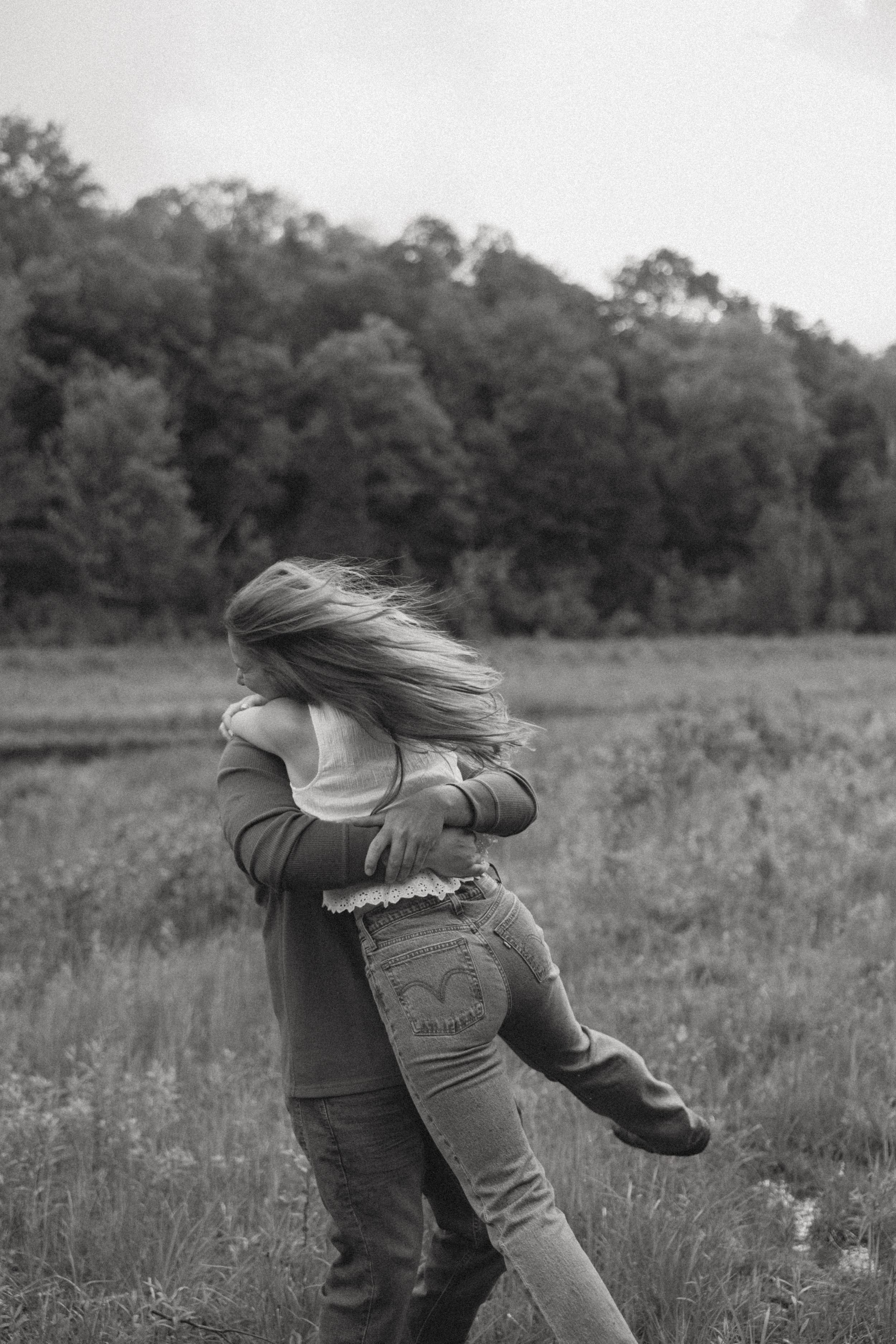 Black and white photo of a couple embracing in a field, woman's hair flowing as man lifts her up.