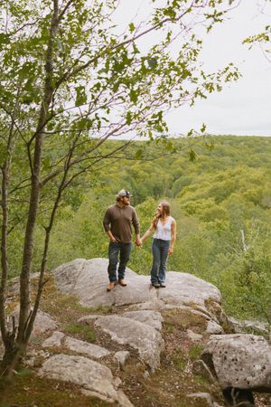 Couple holding hands on rocky overlook surrounded by lush green forest trees.