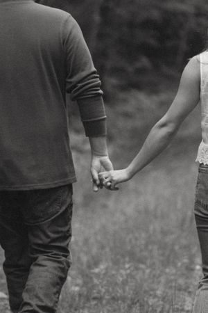 Black and white photo of a couple holding hands while walking through a field, viewed from behind.