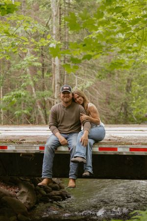 A couple sitting together on a bridge over a stream surrounded by green forest trees.