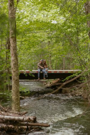 Couple sitting together on a wooden bridge over a creek surrounded by lush green forest trees.