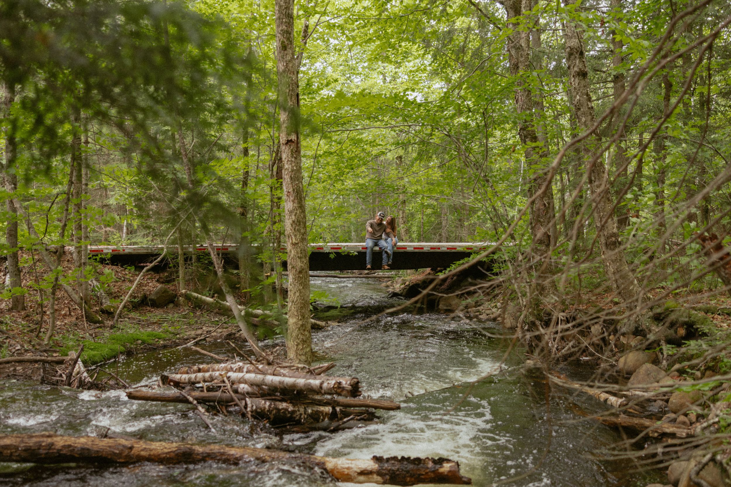 Couple sitting on a wooden bridge over a forest creek surrounded by lush green trees in spring.