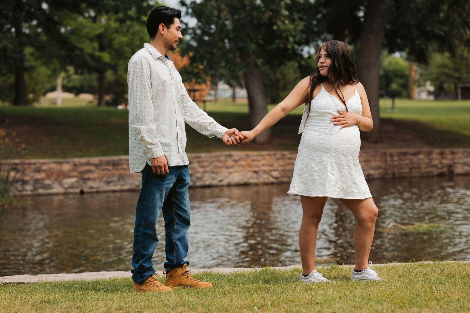 Pregnant couple holding hands by a pond in a park, woman in white dress touching baby bump.