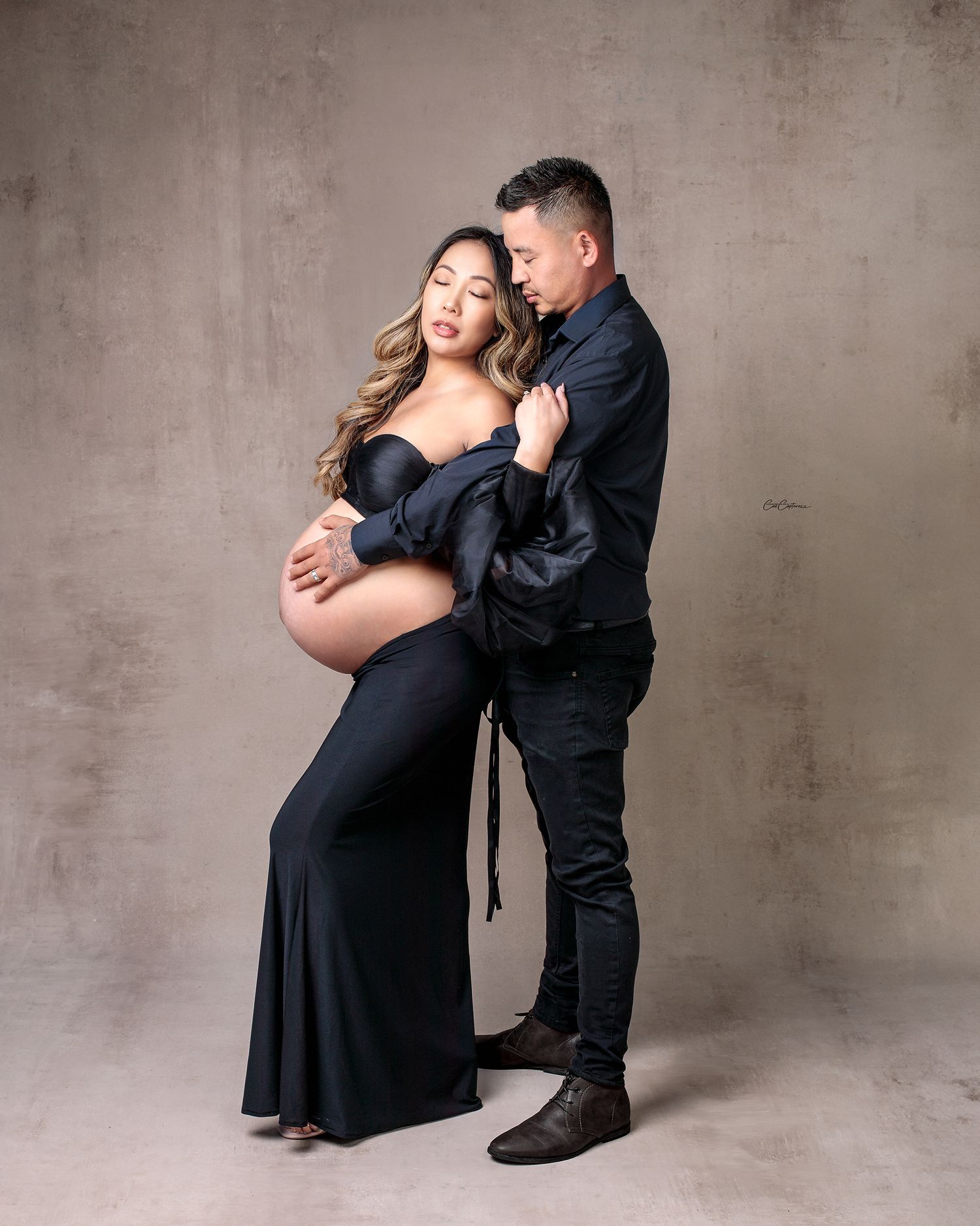 Couples maternity portrait. Man stands behind woman his arms wrapped around her pregnant belly, both eyes closed, woman lovingly touches man