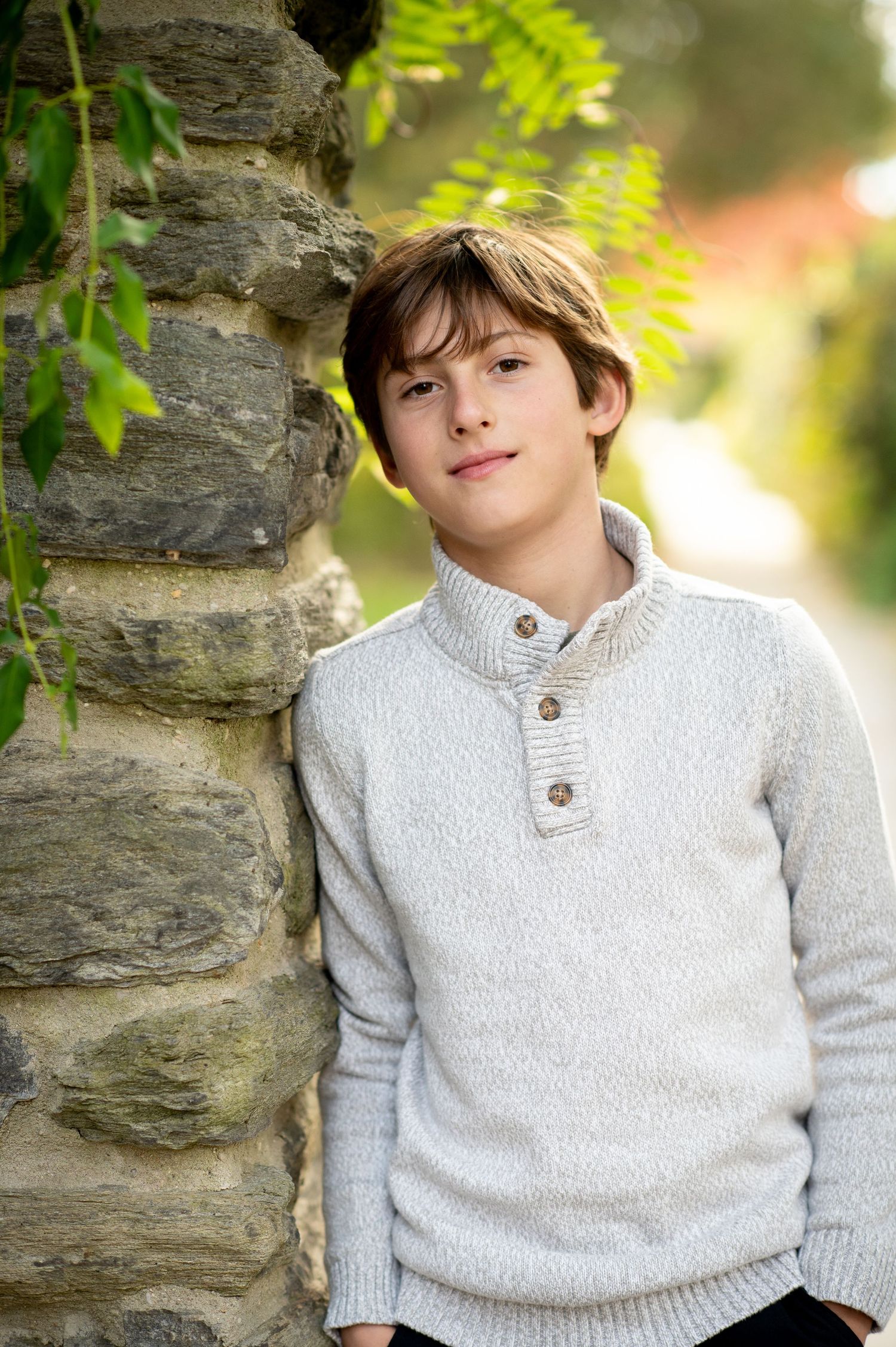A young boy in a white sweater leans against a stone wall outdoors, surrounded by green foliage.