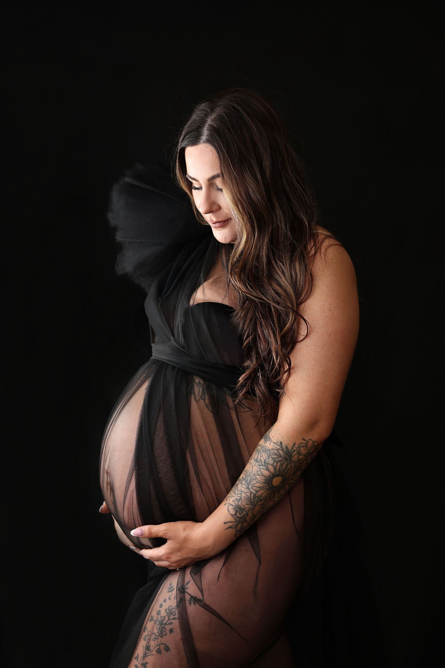 Pregnant woman in sheer black gown cradling baby bump, showcasing tattoos against dark studio background. R.A.M Photography, Stranraer