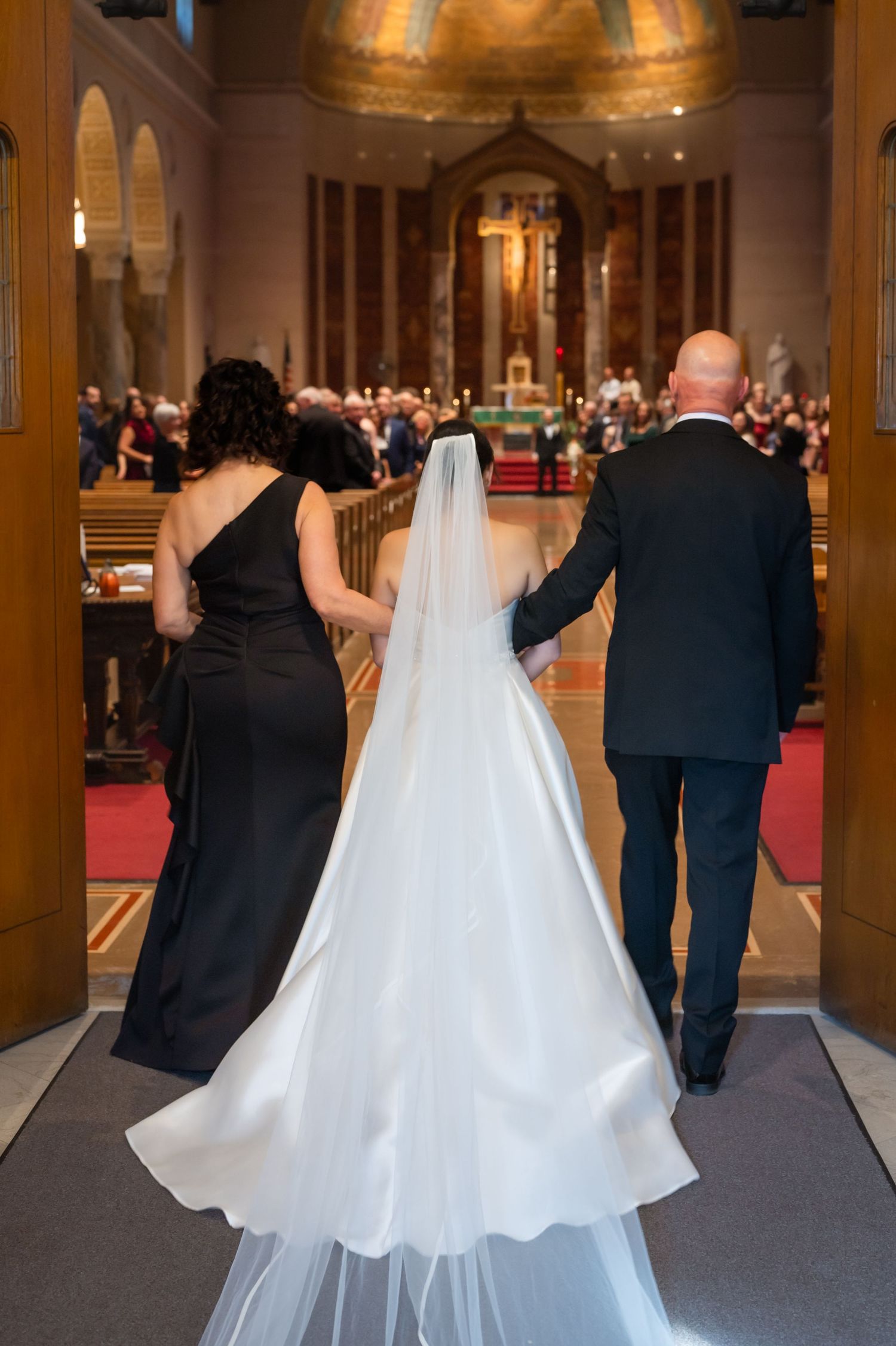 Bride in white gown and veil walks down church aisle flanked by woman in black dress and bald man in suit.