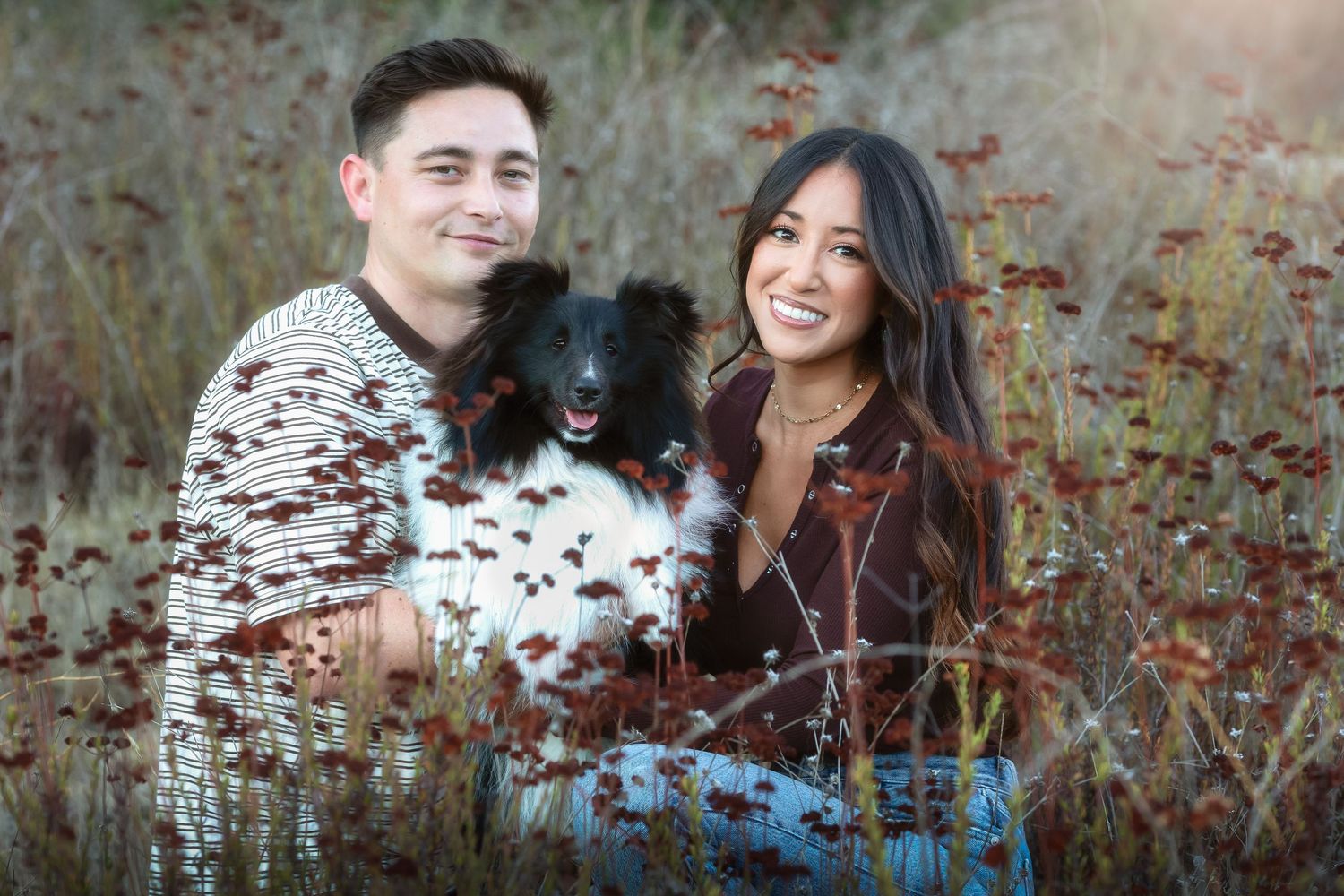 A smiling couple holds a fluffy black and white dog outdoors with green hills in the background.